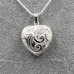 Close-up of a silver heart-shaped pendant with engraved scroll patterns against a gray fabric background, hanging from a chain far above.