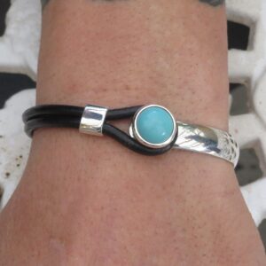 Close-up of a wrist wearing a black leather bracelet with a turquoise circular charm and a silver band.
