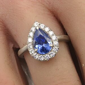 Pear-shaped blue sapphire ring with a diamond halo on a pale metal band, worn on the finger