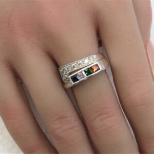 Close-up of a hand wearing a wide silver ring with a row of square-cut colored stones (green, orange, purple, blue) and small surrounding diamonds.